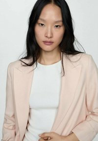 Blazer in soft pink fabric with a tailored design and notched lapels, worn over a white ribbed shirt. Hair is long and dark.