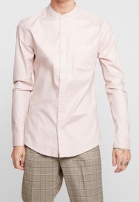 Ljusrosa långärmad skjorta med ståkrage, framficka och full knappslå, tillverkad av mjukt bomullstyg.