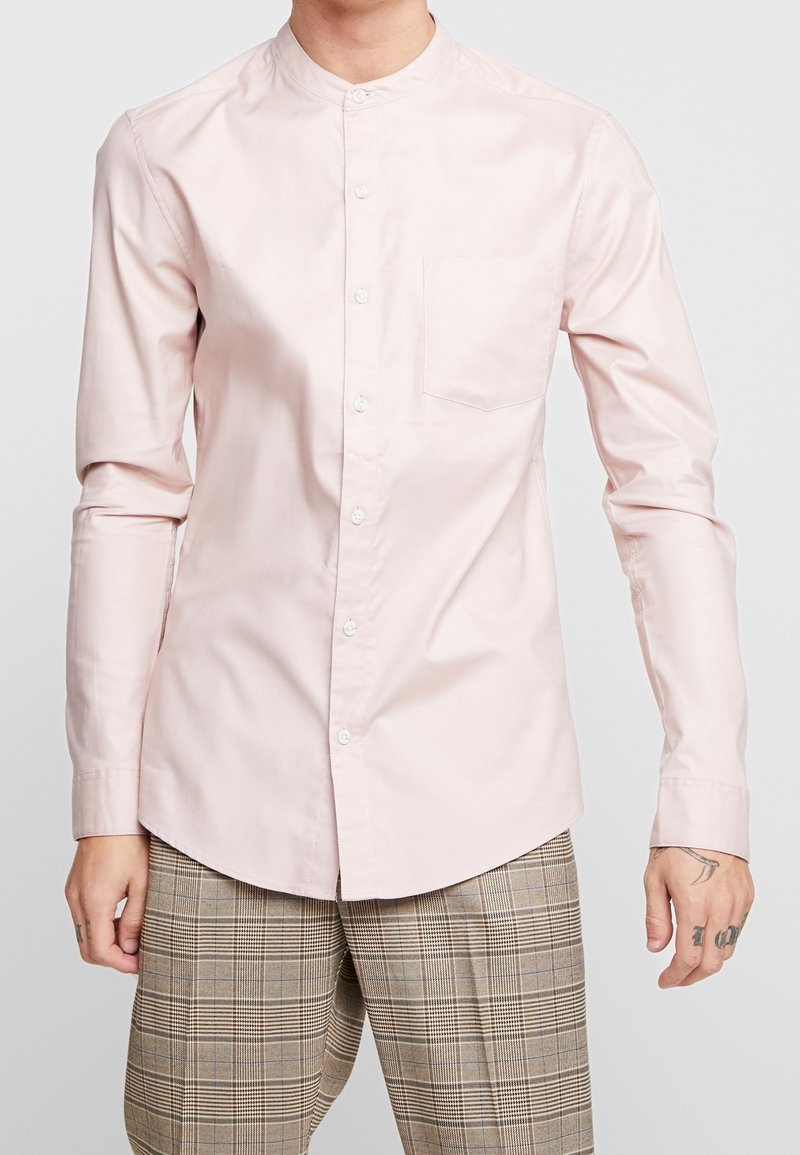Ljusrosa långärmad skjorta med ståkrage, framficka och full knappslå, tillverkad av mjukt bomullstyg.