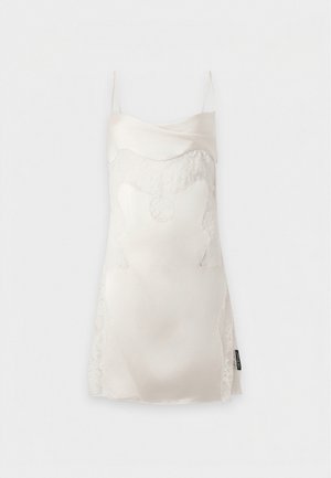 Femme portant une robe nuisette en satin crème sans manches avec des détails en dentelle et des sandales noires à talons bas, debout, une main sur la hanche.