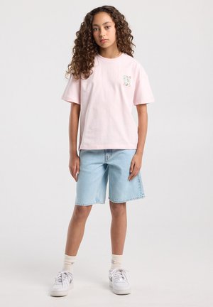 Fille aux cheveux bouclés portant un t-shirt rose clair, un short en denim bleu clair, des chaussettes blanches et des baskets blanches, debout devant un fond uni.
