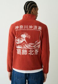 Fonott hajú személy piros polárdzsekit visel, melyen fehér japán hullám művészet és írásjelek láthatók a háton, hozzá kék farmernadrágot hord.