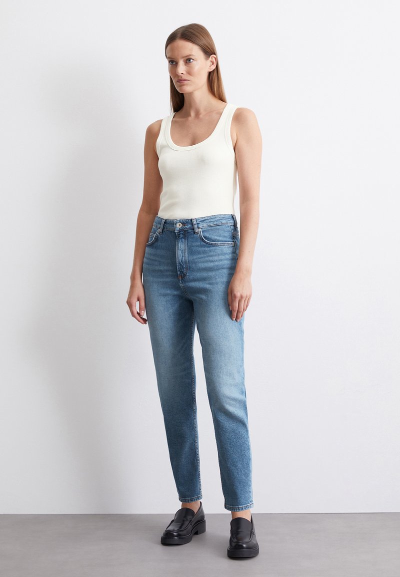 Weißes geripptes Tanktop kombiniert mit hellblauen, gerade geschnittenen Jeans, die über fünf Taschen und eine verwaschene Optik verfügen, ergänzt durch schwarze Loafers.