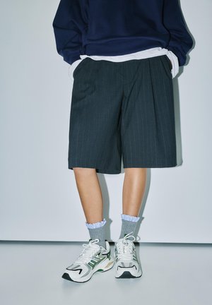 Person trägt einen marineblauen Pullover, ein weißes Unterhemd, dunkle knielange Shorts mit Nadelstreifen, graue gerüschte Socken und weiße Sneaker mit grünen Akzenten.