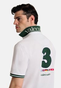 Camisa polo blanca con un collar verde que lleva el texto "valecuatro", rayas verdes en las mangas y un gran "3" en la parte posterior con banderas.