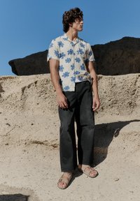 Camisa blanca de manga corta con patrones florales azules, combinada con pantalones negros holgados y sandalias marrones, con un fondo rocoso.