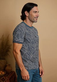 Korte mouwen marineblauw T-shirt met een wit bladenpatroon, ronde hals met teal rand en een enkele zak op de borst. Gedragen met blauwe jeans.