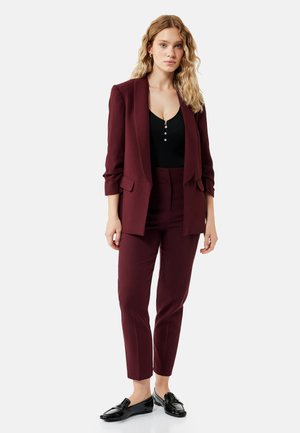 Kvinde med bølget blondt hår står iført en burgunderrød dragt med skræddersyede bukser, en blazer, sort top og sorte loafers på hvid baggrund.