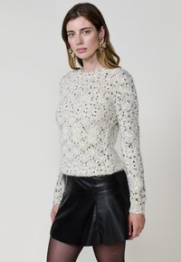 Weißer gestrickter Pullover mit schwarzen und gelben Punkten, lange Ärmel, runder Hals; kombiniert mit einem schwarzen Leder-Minirock.