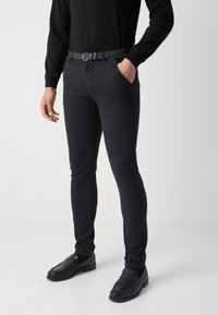 Pantaloni neri aderenti realizzati in un tessuto liscio, abbinati a una cintura nera e mocassini scuri, su uno sfondo neutro con una maglia nera coordinata.