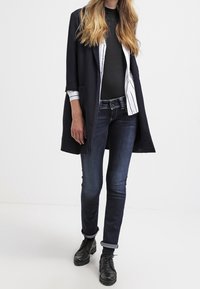 Lång, marinblå kappa över en svart topp, vitrandig skjorta och mörk denimjeans. Svarta snörskor med uppvikta byxben.