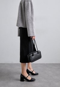 Mustat Mary Jane -korot, joissa on paksu korko, yhdistetty harmaaseen ribbineulepaitaan ja mustaan polvipituiseen hameeseen. Pienessä mustassa käsilaukussa pidetään yhdessä kädessä.