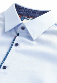 Hele sinine polo särk, millel on tekstuuriga kangas, nööbiga sulgemine ning sinise ja lillemustriga kaeluse vooder.