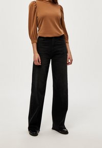 Kvinna som bär en brun puffärmad topp, högmidjade svarta vida jeans och svarta glänsande loafers mot en enkel vit bakgrund.