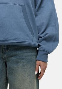 Blue cotton sweatshirt with a kangaroo pocket and ribbed cuffs, paired with faded denim jeans, showing a relaxed fit.