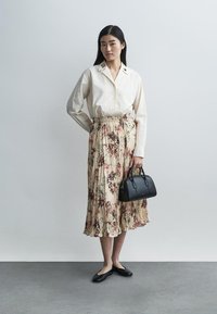 Chemise crème boutonnée avec col brodé, jupe plissée fleurie beige, sac à main noir et ballerines noires. Texture lisse, design fluide.