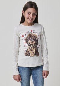 Ein junges Mädchen mit langen braunen Haaren trägt ein weißes Langarmshirt, das einen lockigen Hund mit Sonnenbrille und Herzen zeigt, kombiniert mit blauen Jeans.