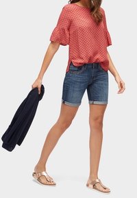 Punakuvioinen pusero, jossa on lyhyet röyhelöhihat, yhdistetty farkkushortseihin ja ruskeisiin sandaaleihin. Kädessä tummansininen neulepaita.