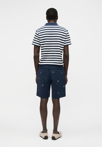 Hombre que lleva pantalones cortos azul marino bordados y una camiseta polo de manga corta a rayas blanca y azul marino, visto desde atrás, sobre un fondo blanco.