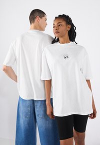 Hvid oversized t-shirt med korte ærmer og et trykt "hell" logo på fronten. Modellen parrer den med denim shorts og sorte cykelshorts.