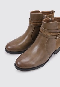 Botas de tobillo marrones con un acabado de cuero brillante, que cuentan con una correa texturizada con tachuelas y un cierre de cremallera lateral para ponerlas y quitarlas fácilmente. Suela plana.