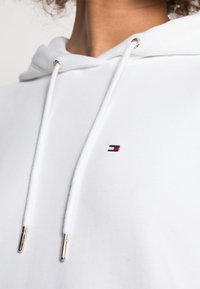 Biała bluza z kapturem z przednią kieszenią kangurką, ze sznurkami zakończonymi srebrnymi metalowymi końcówkami oraz małym haftowanym logo w kolorze czerwonym, białym i niebieskim.