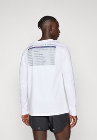 Man with braided hair wears white long-sleeve shirt with Berlin departures table print and black shorts, facing away on neutral background.