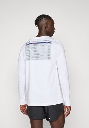 Man with braided hair wears white long-sleeve shirt with Berlin departures table print and black shorts, facing away on neutral background.