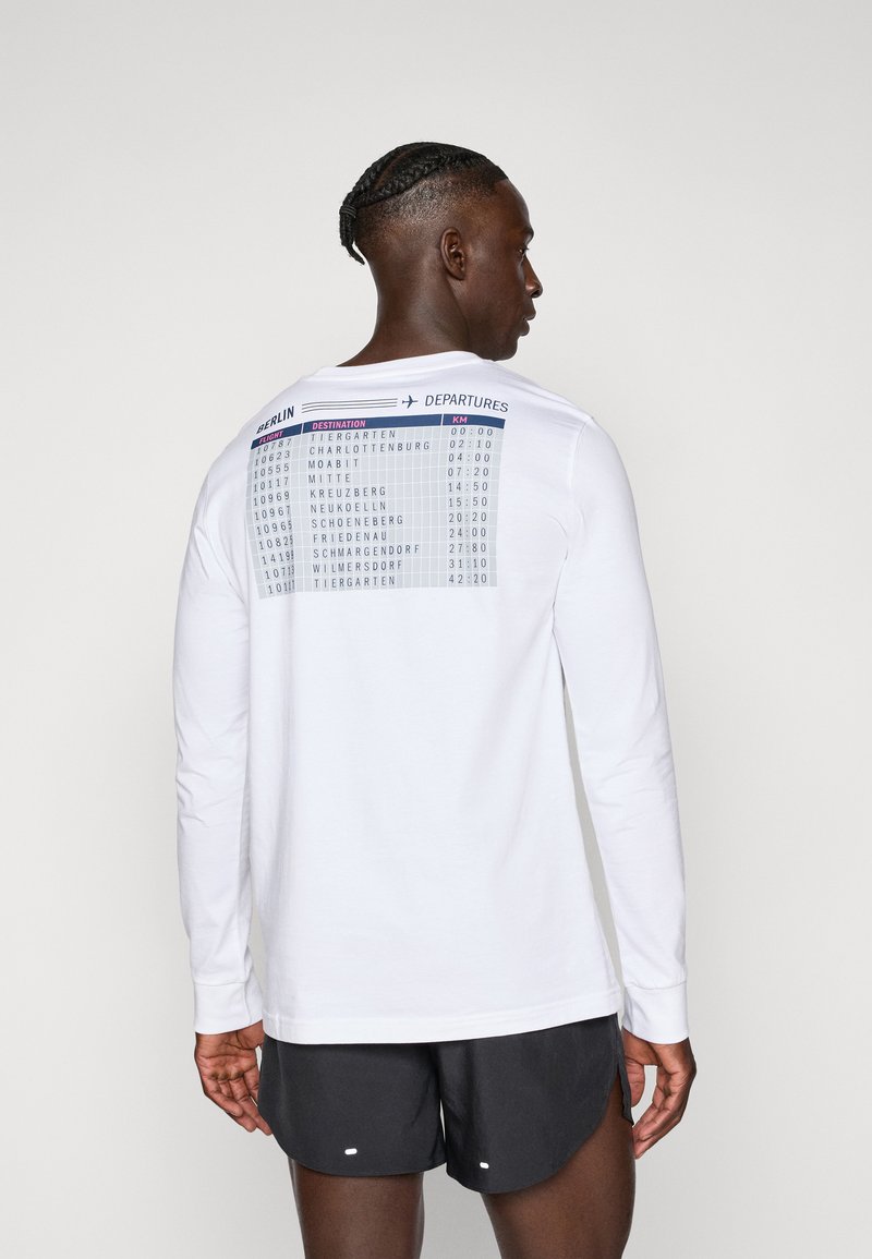 Homme avec des cheveux tressés portant une chemise blanche à manches longues avec un imprimé de tableau des départs de Berlin et un short noir, de dos sur un fond neutre.