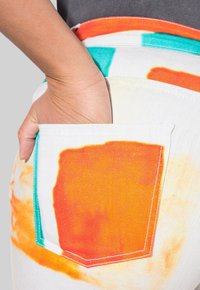 Los jeans de denim blanco presentan un diseño colorido en los bolsillos con acentos en naranja y turquesa. La tela parece texturizada y robusta.