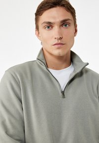 Jersey de color verde oliva claro hecho de una tela suave y texturada. Presenta un cuello alto con cierre de cremallera y una capa interior blanca visible.
