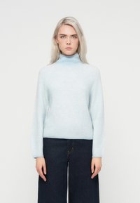 Gai zilas turtlenecks džemperis, kas izgatavots no mīkstas adīta materiāla, ar brīvu piegriezumu un garām piedurknēm, kombinēts ar tumšiem platām kājām džinsiem.