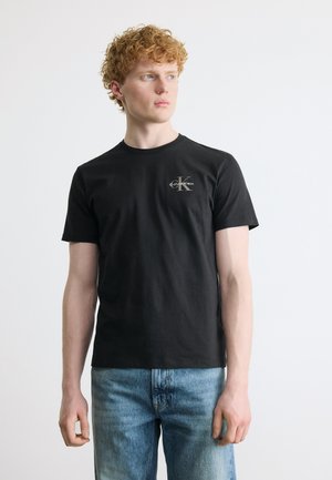 Fiatal férfi göndör vörös hajjal, fekete Calvin Klein pólót és kék farmert visel, balra néz egy egyszerű fehér háttér előtt.