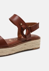 Sandale compensée en cuir marron avec semelle en jute tressée, bride de cheville avec une boucle circulaire foncée et un clou doré.