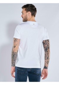 White cotton t-shirt with a crew neck, short sleeves, and a small logo detail at the back. Model wears dark jeans, showing detailed tattoos.