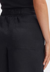 Un pantalon en lin noir avec une taille élastique et une poche arrière. Le tissu a une apparence texturée et une coupe décontractée.