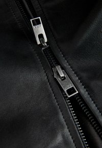 Közeli kép egy fekete bőrdzseki cipzárjáról, amely ezüstszínű fém húzófülekkel részlegesen lefelé van húzva a ruhadarabon.