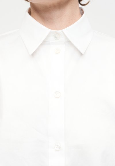 Κοντινό πλάνο ενός ατόμου που φοράει λευκό πουκάμισο με γιακά και κουμπιά, με ορατά τα κουμπιά και μέρος του λαιμού και του πηγουνιού.