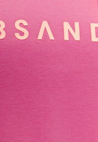 Top senza maniche di colore rosa con una texture morbida, caratterizzato da testo in rilievo di un rosa chiaro che recita "B SAND." Il tessuto è liscio e uniforme.