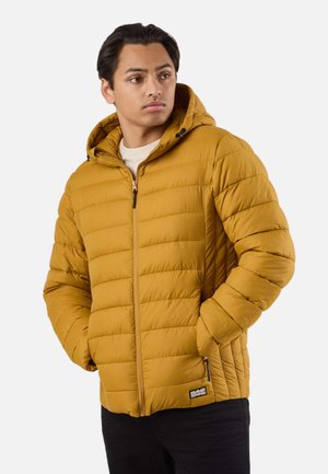Young man wearing mustard yellow quilted hooded jacket with hands in pockets, looking slightly to the side.
