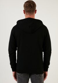 Hoodie nero con texture, con cappuccio regolabile tramite lacci, polsini e orlo a coste, visto da dietro. Realizzato in tessuto morbido e elasticizzato.