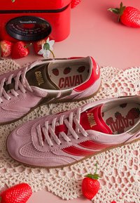 Paire de baskets en suède rose et rouge avec la marque « CAVAL », semelles intérieures à motif fraise, à côté d'une balance rouge et de fraises artificielles sur un tapis en dentelle.
