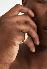 Guldring med et glat, buet design, båret på en hånd med tekstureret, mørk hud, der fremhæver ringens form og metalliske finish.