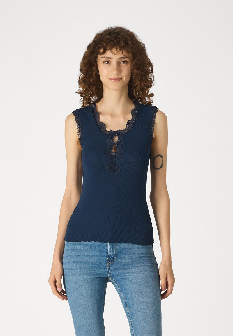 Ναυτικό μπλε αμάνικο τοπ με δαντελένια διακόσμηση, στρογγυλή λαιμόκοψη και λεπτομέρεια με κλειστό λαιμό μπροστά. Ελαφρώς εφαρμοστή γραμμή με υφασμένο ριχτό ύφασμα.