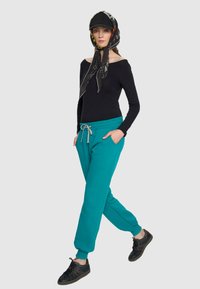 Turkusowe spodnie dresowe z bocznymi kieszeniami i szarym sznurkiem, zestawione z czarną bluzką bez ramiączek. Modelka nosi czapkę w kolorze czarnym i wzorzystą chustę.