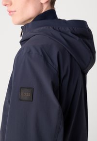 Marineblauwe hooded jacket van gladde stof, met een zwart rechthoekig label met het "BOSS" logo op de linker mouw.