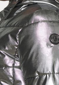 Veste matelassée argent métallique brillante en gros plan, avec des coutures matelassées et un patch logo rond noir sur la manche.