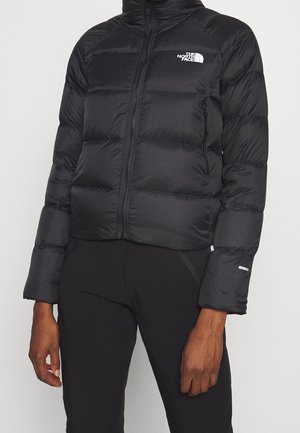 Person iført en sort dunjakke med lynlås foran, The North Face-logo på brystet og "550" mærkat på venstre ærme, kombineret med sorte bukser.