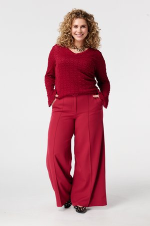 Femme souriante aux cheveux bouclés portant un haut rouge texturé à manches longues, un pantalon large rouge, un collier et des chaussures noires cloutées, les mains dans les poches.