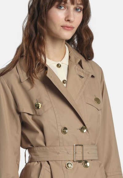 Beige trench coat with a double-breasted design, gold buttons, belted waist, and two chest pockets. Smooth texture with tailored fit.